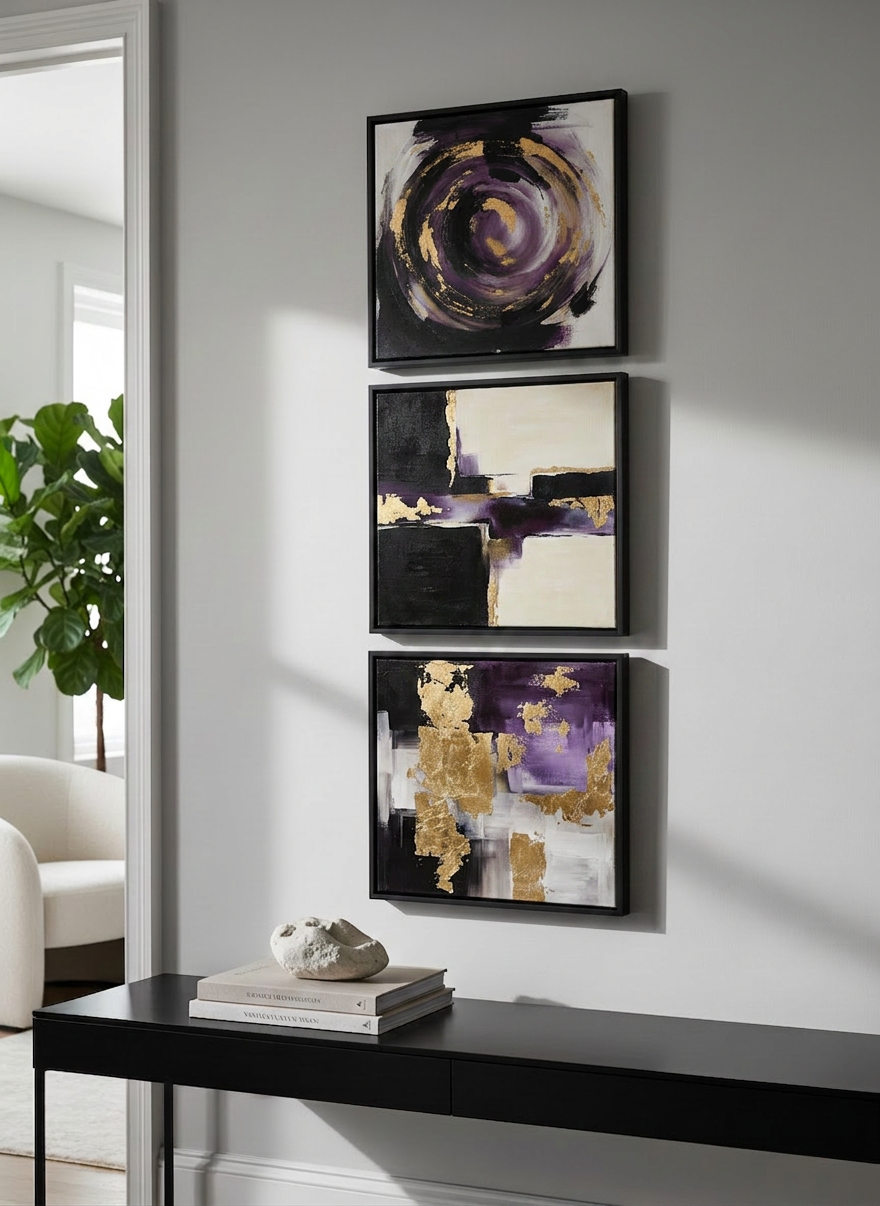 A sophisticated interior vignette showcasing a trio of custom expressionist canvases hung in a vertical arrangement on a smooth, dove-grey wall. Each painting features bold, gestural brushstrokes in a restrained palette of ink black, warm taupe, and smoky blue, with hints of muted gold leaf catching the light. Beneath them, a sleek black console holds stacked design books and a single sculptural stone object. Soft, diffused natural light from an unseen window creates gentle, elongated shadows and lightly illuminates the textured paint ridges. Captured in photographic realism from a slightly angled perspective, with sharp focus on the artwork and a subtly blurred background. The atmosphere is refined, modern, and curated, emphasizing the role of bespoke abstract art as a key element of interior design.